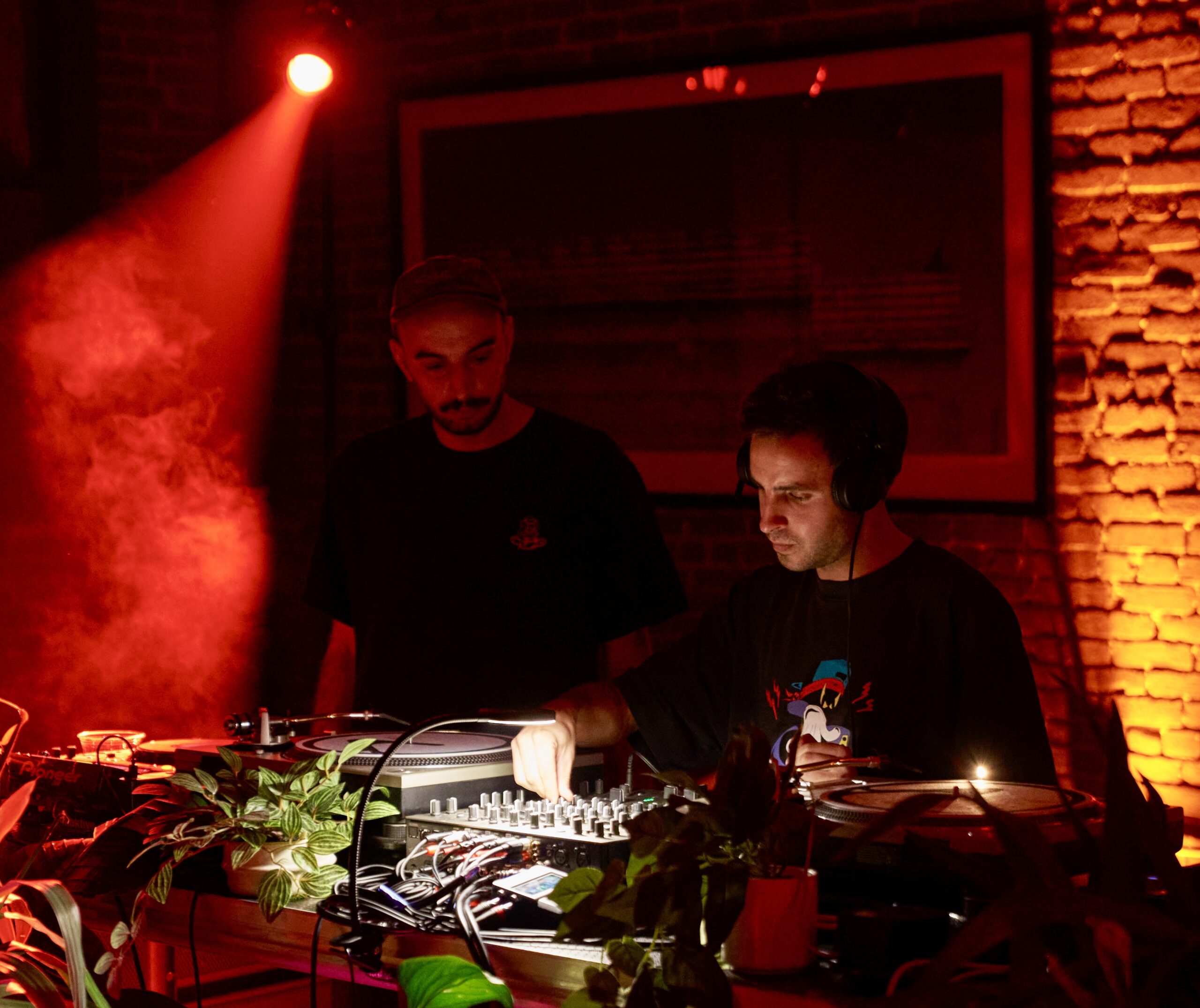 Two DJs perform in a dimly lit room with red lighting and smoke. One operates a mixer while the other stands beside turntables. Potted plants are in the foreground.